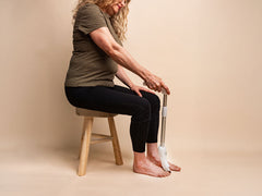 Woman sitting on a stool holding a white long handled nail filing against a beige background